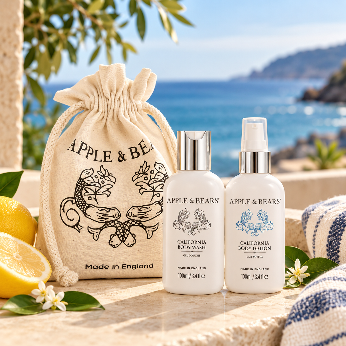 APPLE & BEARS California Travel Toiletries Set featuring 100ml Body Wash and 100ml Body Lotion in Bergamot Waters fragrance, displayed alongside a reusable cotton gift bag on a sunlit stone terrace overlooking the sea.
