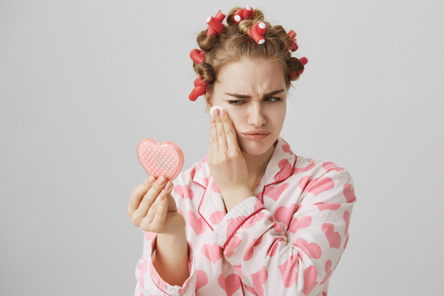 A person with pink hair curlers in their hair gently cleansing their face with a round cotton pad while checking their skin blemishes in a mirror reflection.