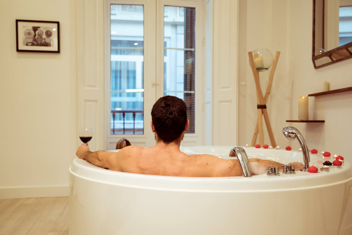 A man relaxing in a luxurious, warm ambient bubble bath interior setting enjoying a glass of red wine during a premium self-care routine.