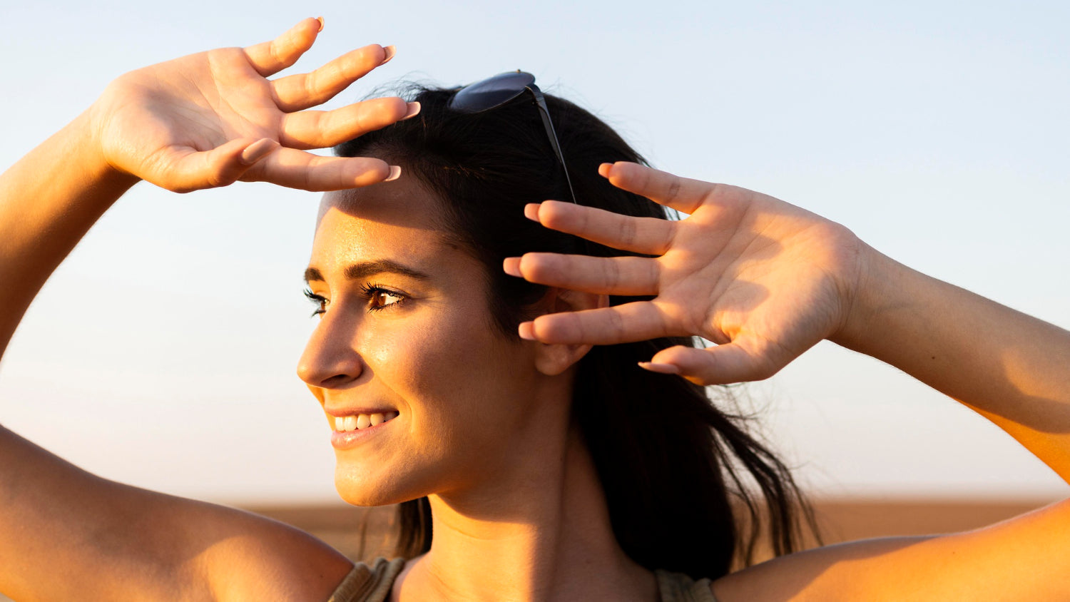 A smiling woman with sunglasses on her head shielding her eyes from bright sunlight during golden hour.