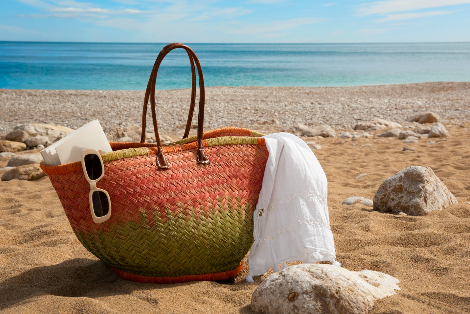 A stylish orange and green woven beach tote bag resting on a sandy pebble shore next to a white linen wrap towel, sunglasses, and Apple and Bears luxury eco-friendly travel skincare sets.