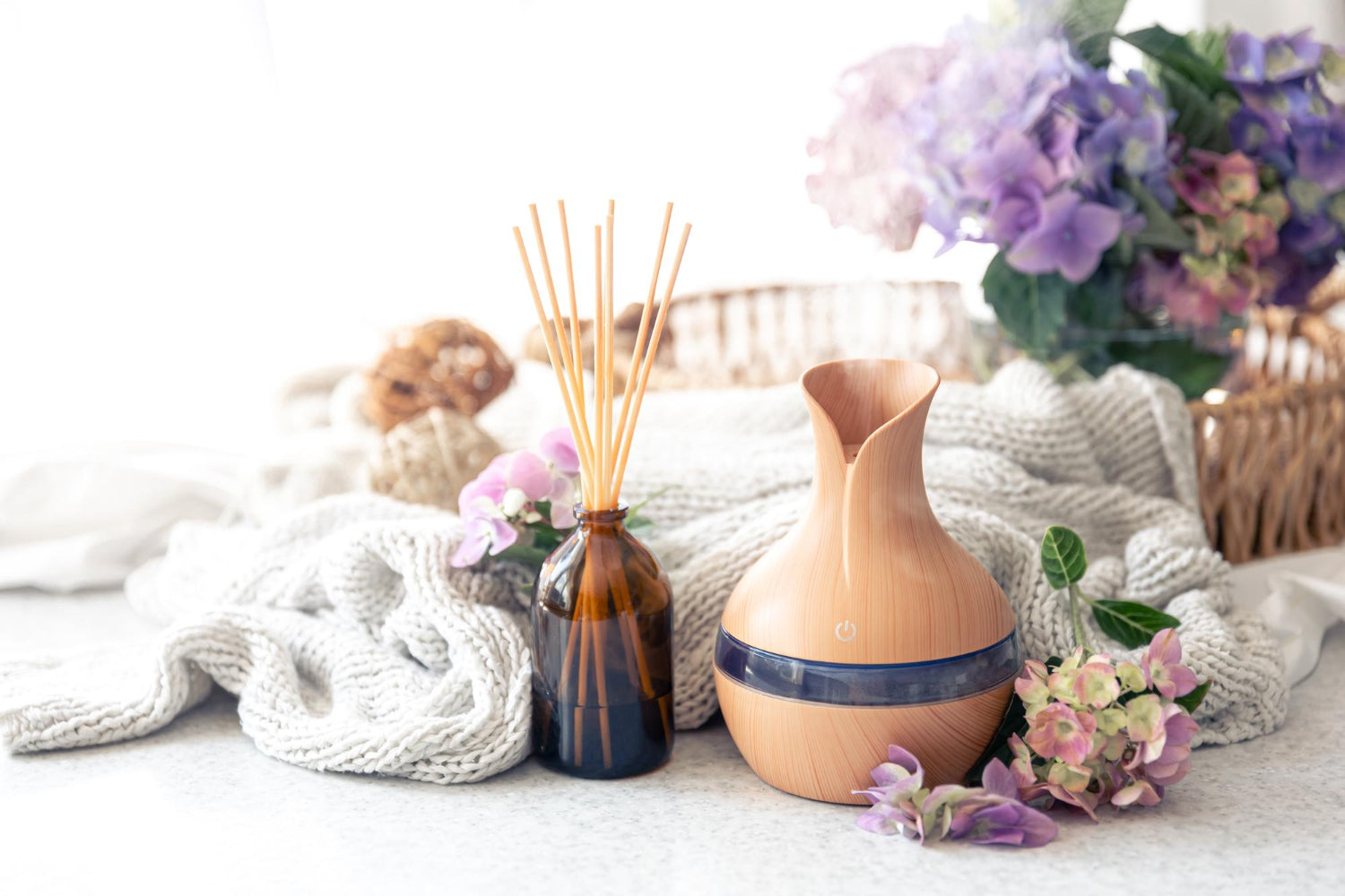 An elegant wooden electronic aromatherapy mist diffuser operating on a cozy knit blanket next to a clear glass vial of essential oils and purple lavender flowers.