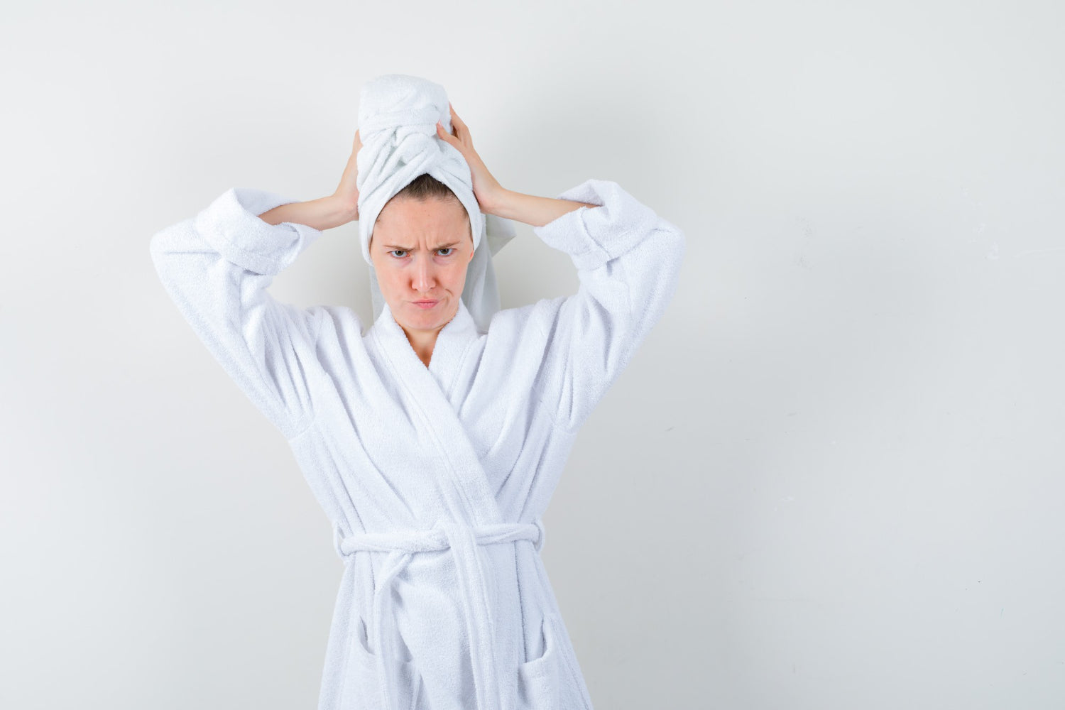 A frustrated woman in a premium white luxury spa robe with a white wash towel wrapped around her hair, inspecting her sensitive skin texture in a bright setting.