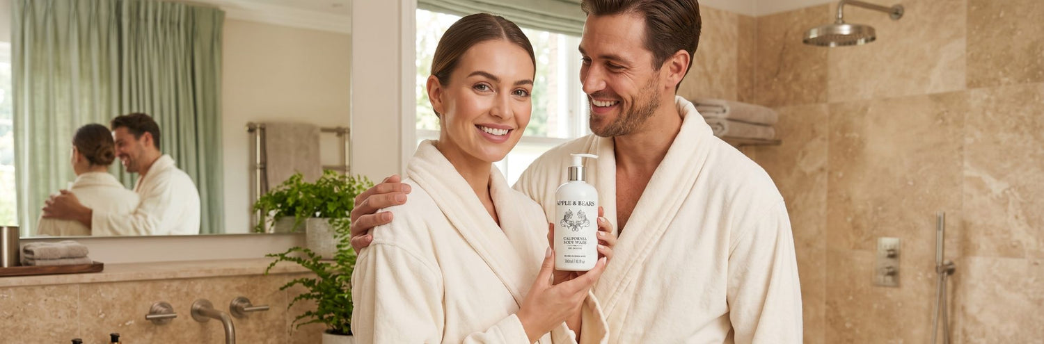 A smiling couple in white spa robes holding a premium bottle of Apple and Bears California organic citrus body wash together in an elegant, sunlit bathroom.