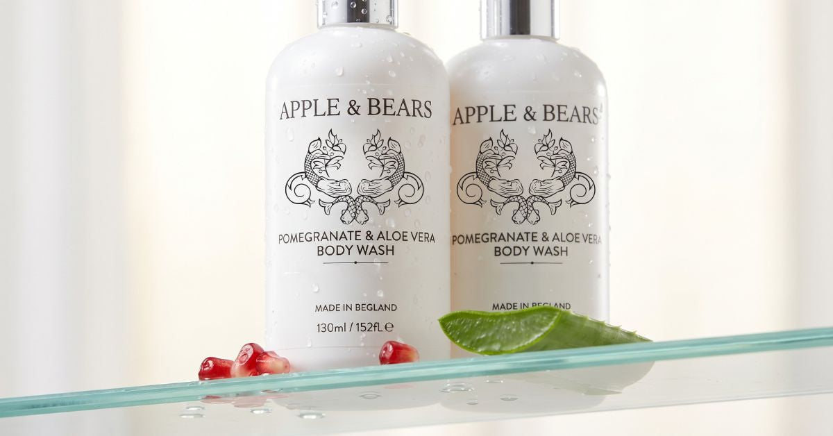 Luxury eco-friendly body wash bottles from Apple and Bears styled behind a modern clear glass shelf with organic botanical ingredients including fresh aloe vera slices and whole pomegranates.