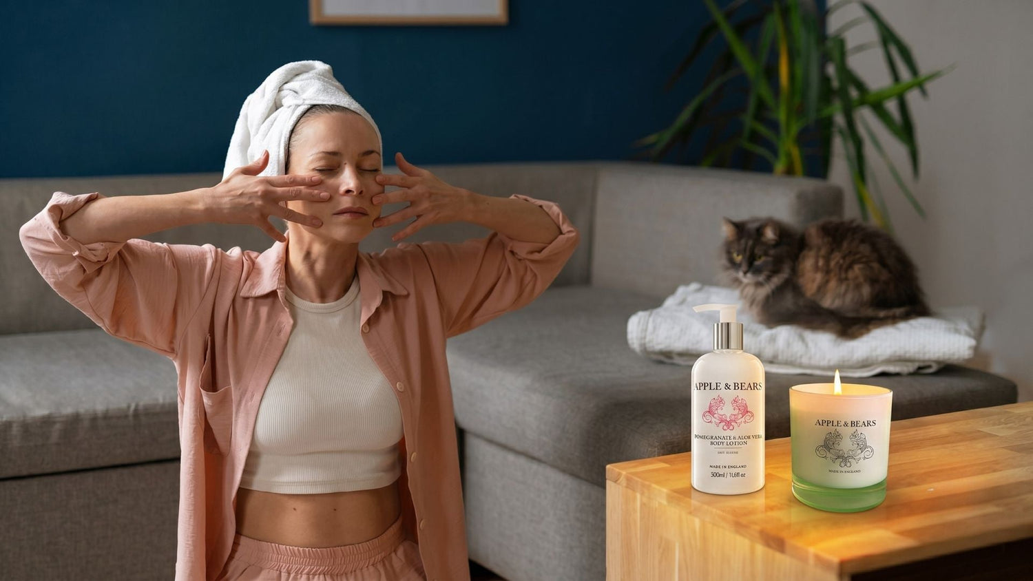 A serene individual with a white hair towel gently massaging their face in a beautifully styled bathroom setting next to an Apple and Bears luxury body care bottle and lit ambient candles.