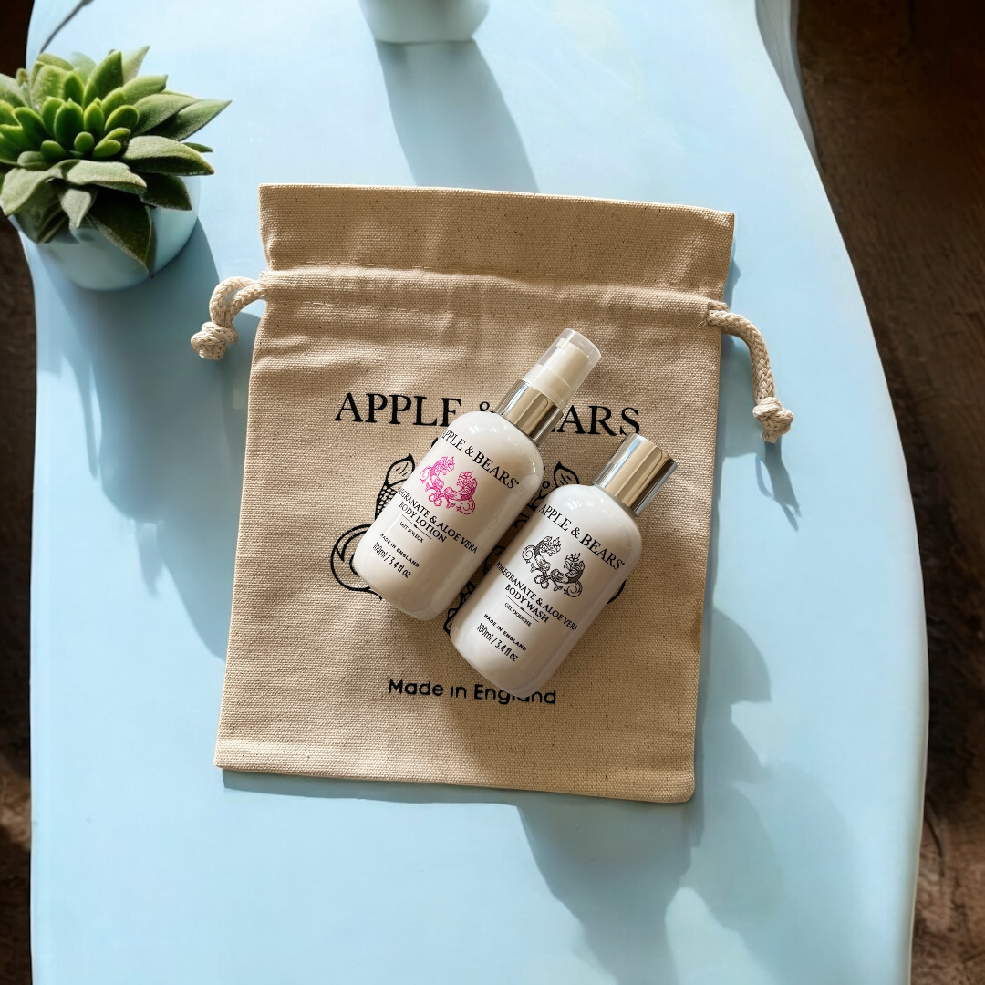 An overhead flat-lay of Apple and Bears certified organic Pomegranate and Aloe Vera travel size body wash and body lotion bottles styled on an organic canvas drawstring pouch over a clear blue surface.