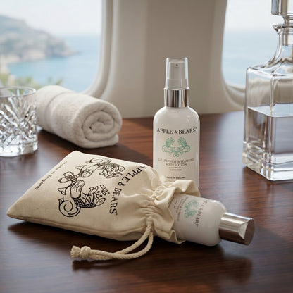 Apple &amp; Bears Grapefruit and Seaweed Body Lotion 100ml with eco cotton drawstring bag on dark wood table with sea view, rolled towel and crystal glassware