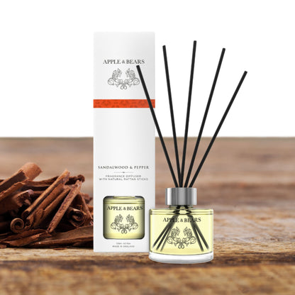 Lifestyle display of the Apple &amp; Bears Sandalwood &amp; Pepper Reed Diffuser set on a rustic wooden surface with natural sandalwood bark accents, highlighting the elegant 125ml bottle and black rattan reeds.