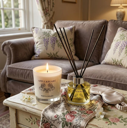 Apple &amp; Bears Jasmine Home Fragrance Gift Set with Soy Wax Candle burning and Reed Diffuser on a vintage painted coffee table with silk sleep mask, jasmine flowers and floral rose cushions in a cosy English cottage living room, Made in England