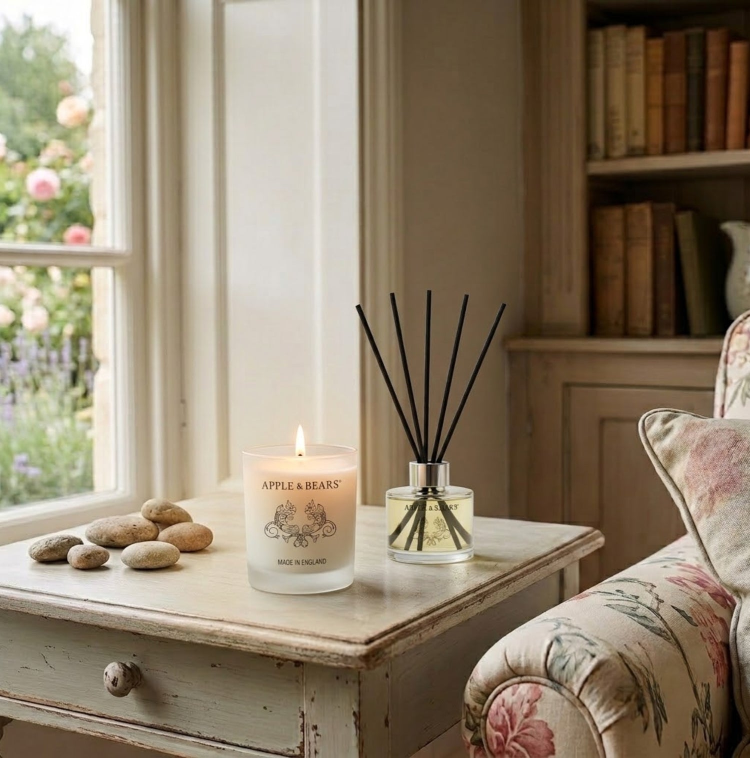 Apple &amp; Bears Lavender Home Fragrance Gift Set with Soy Wax Candle burning and Reed Diffuser on a painted vintage side table in a cosy English cottage living room with floral armchair, pebbles and rose garden views, Made in England