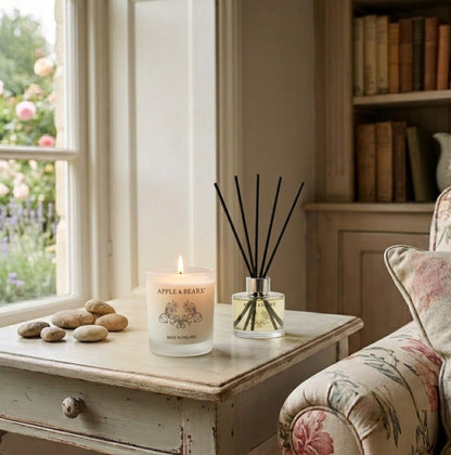 Apple &amp; Bears Lavender Home Fragrance Gift Set with Soy Wax Candle burning and Reed Diffuser on a painted vintage side table in a cosy English cottage living room with floral armchair, pebbles and rose garden views, Made in England
