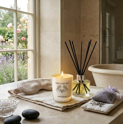 Apple &amp; Bears Lavender Home Fragrance Gift Set with Soy Wax Candle burning and Reed Diffuser on a stone spa bathroom surface with silk sleep mask, lavender sachet, hot stones and English lavender garden views, Made in England