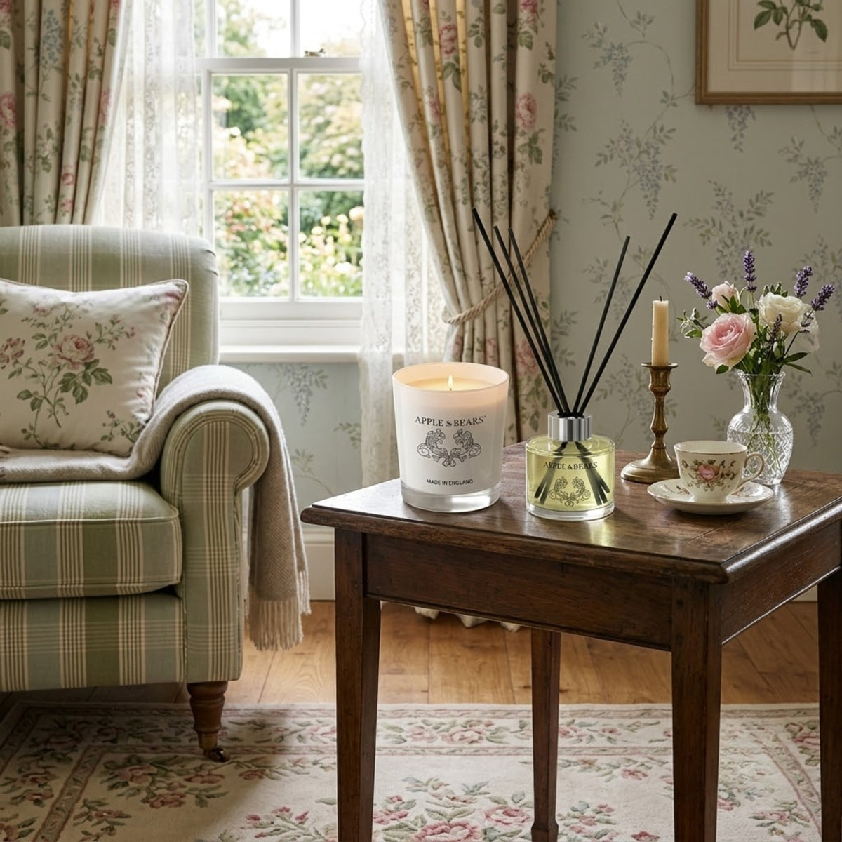 Apple &amp; Bears Sandalwood &amp; Pepper Home Fragrance Gift Set with Soy Wax Candle burning and Reed Diffuser on a dark oak side table with English bone china teacup, brass candlestick, fresh pink roses and lavender in a cosy English cottage living room with floral wallpaper and garden views, Made in England