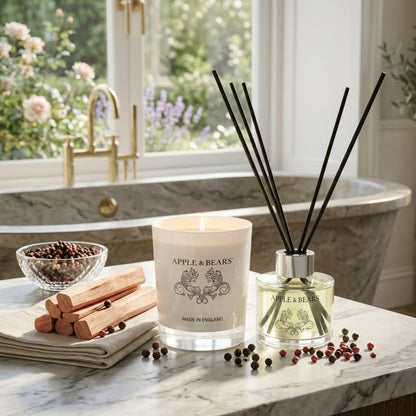 Apple &amp; Bears Sandalwood &amp; Pepper Home Fragrance Gift Set with Soy Wax Candle burning and Reed Diffuser on a marble bathroom surface with sandalwood sticks, pink and black peppercorns and English rose garden views through a gold tap window, Made in England