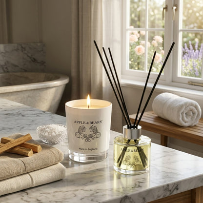 Apple &amp; Bears Sandalwood &amp; Pepper Home Fragrance Gift Set with Soy Wax Candle burning and Reed Diffuser on a marble spa bathroom surface with sandalwood sticks, bath salts, rolled white towel and English rose and lavender garden views through a sash window, Made in England