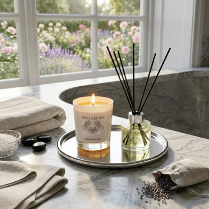 Apple &amp; Bears Sandalwood &amp; Pepper Home Fragrance Gift Set with Soy Wax Candle burning and Reed Diffuser on a silver tray on a marble spa surface with hot stones, bath salts, linen towels, lavender sachet and English rose and lavender garden views, Made in England