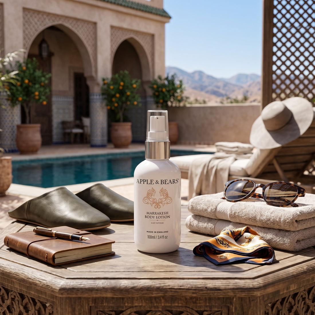 Apple &amp; Bears Marrakesh Body Lotion 100ml travel size on wooden table by a Moroccan riad pool with leather babouche slippers, sunglasses, journal and Atlas Mountains view